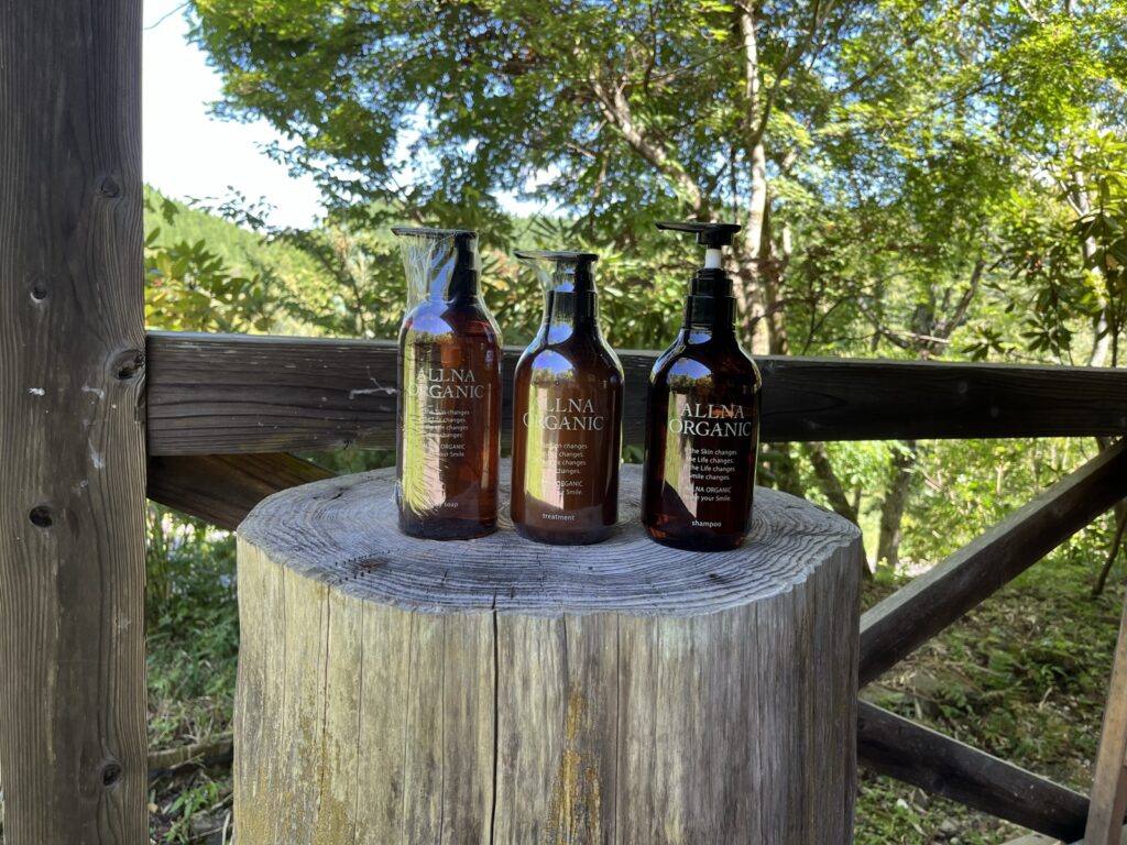 切り株の上に並べた ALLNA ORGANIC のボディソープ・トリートメント・シャンプーの3本。背景は阿蘇の森