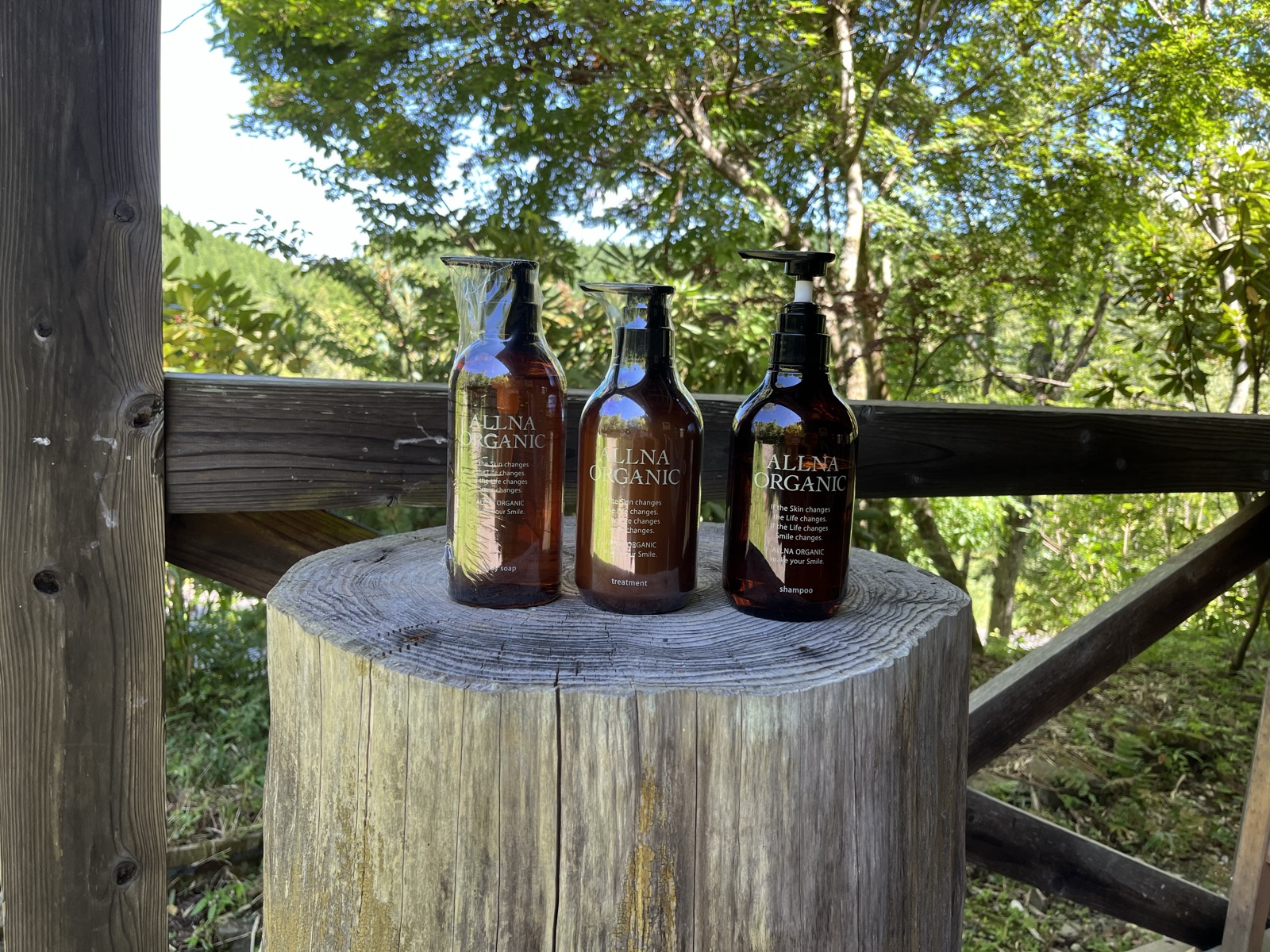 切り株の上に並べた ALLNA ORGANIC のボディソープ・トリートメント・シャンプーの3本。背景は阿蘇の森