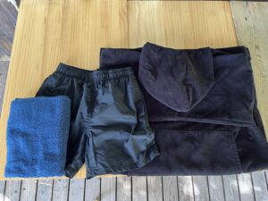 木のテーブルに広げたミキタオサウナのレンタル品。左からたたんだ黒いフェイスタオル、ウエストがゴムの黒い男性用水着、フード付きの黒いタオル地のポンチョ、奥に黒いバスタオルが並んでいる